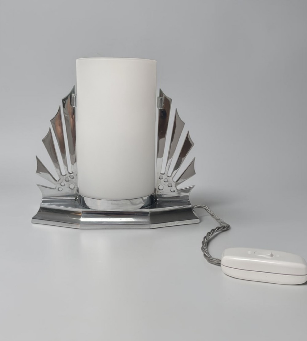 An Art Deco chrome table lamp featuring a cylindrical frosted glass shade flanked by geometric sunburst wings on a stepped chrome base.