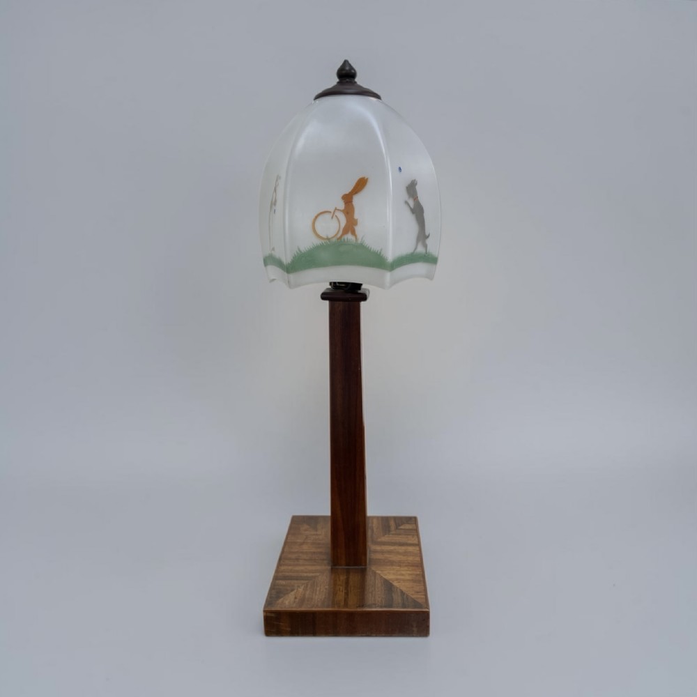 An Art Deco table lamp featuring a square parquetry wooden base, a tall central column stem, and a frosted glass shade decorated with painted illustrations of animals.