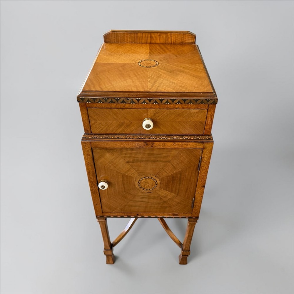 An Art Deco warm brown walnut bedside table featuring geometric marquetry inlay, a single drawer above a cupboard door, and curved lower stretchers.