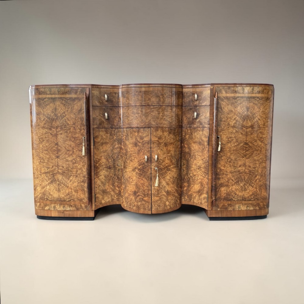 An Art Deco burr walnut sideboard featuring a contoured front, book-matched timber veneers, central double doors, flanking side cupboards, and metallic handles.