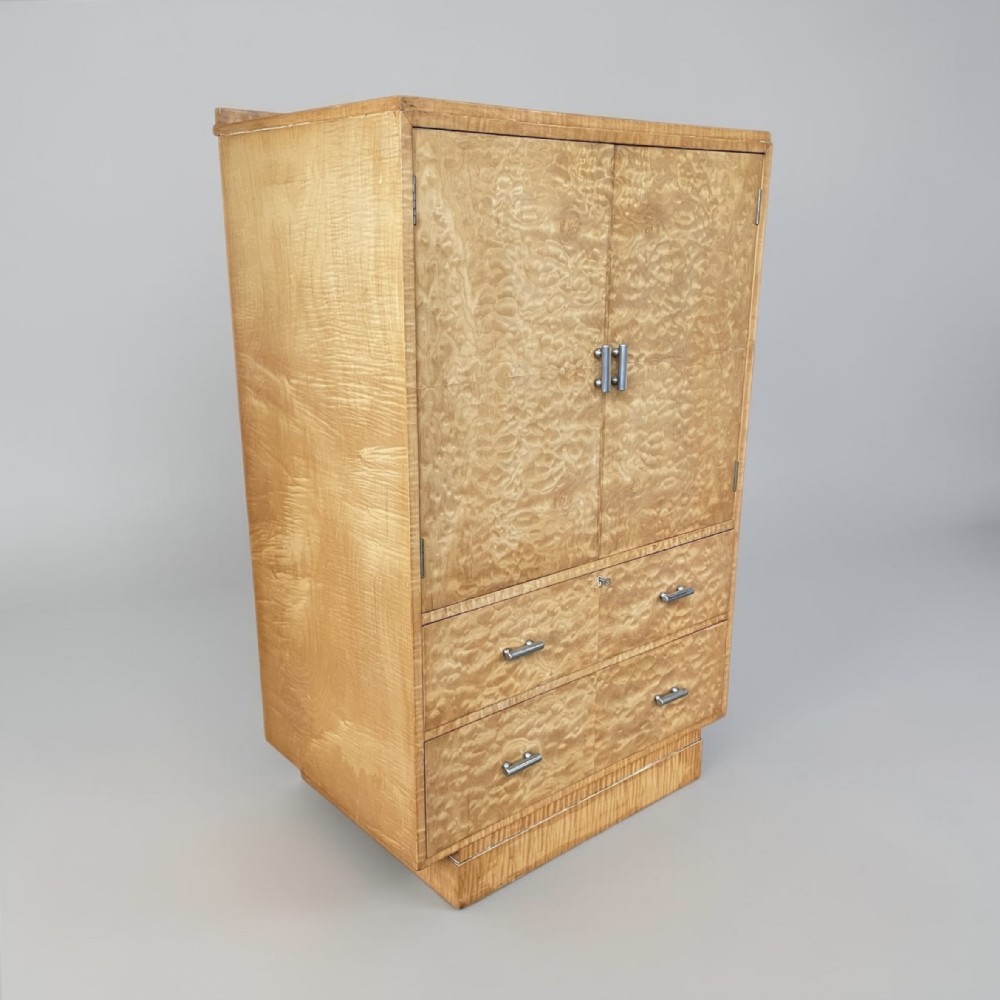 An Art Deco blonde maple tallboy featuring a double-door top cupboard above two lower drawers on a plinth base.