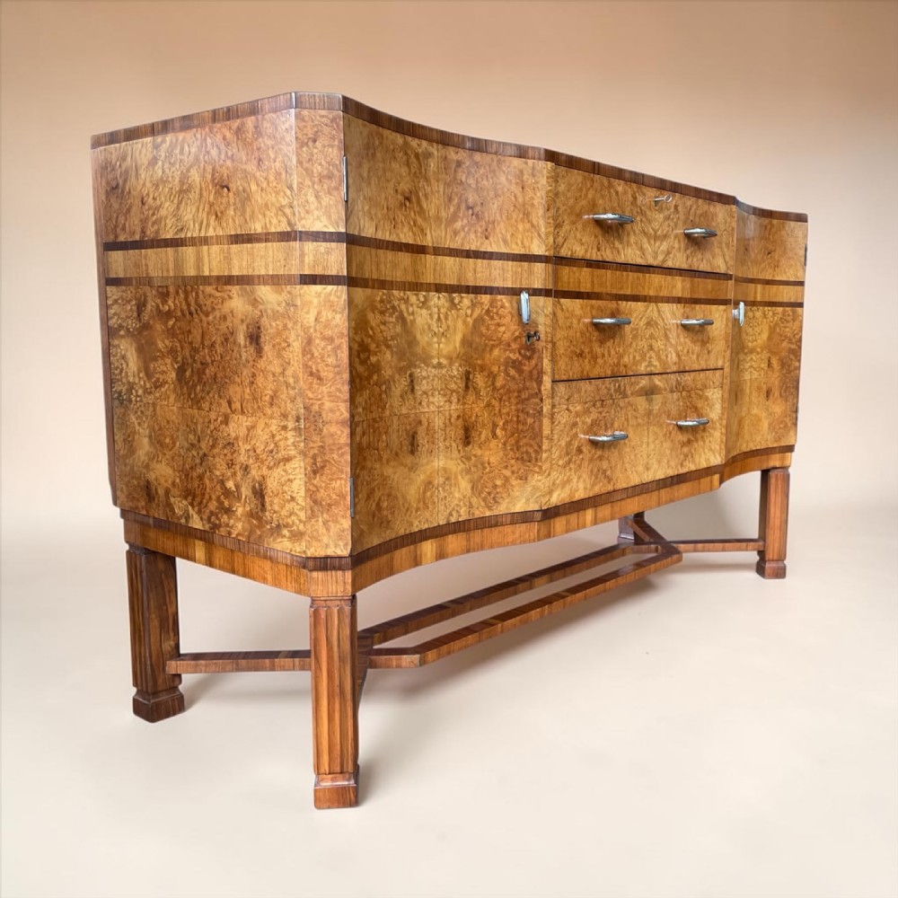An Art Deco burr walnut sideboard featuring a curved serpentine front, three central drawers with metal handles, side cupboards, and fluted legs joined by a stretcher.