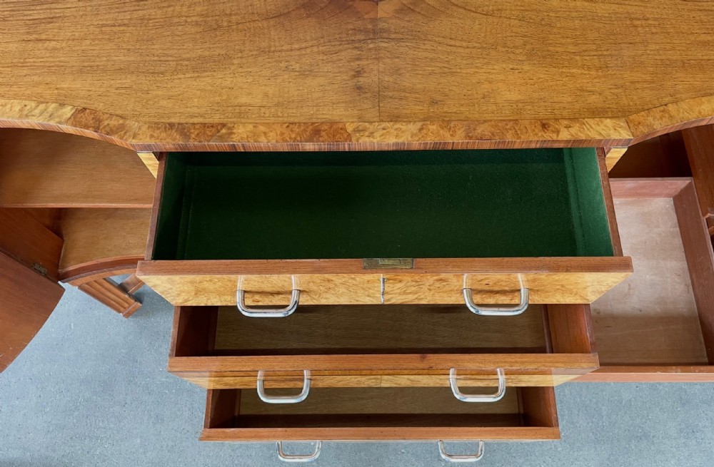 An Art Deco burr walnut sideboard featuring a curved serpentine front, three central drawers with metal handles, side cupboards, and fluted legs joined by a stretcher. View VIII