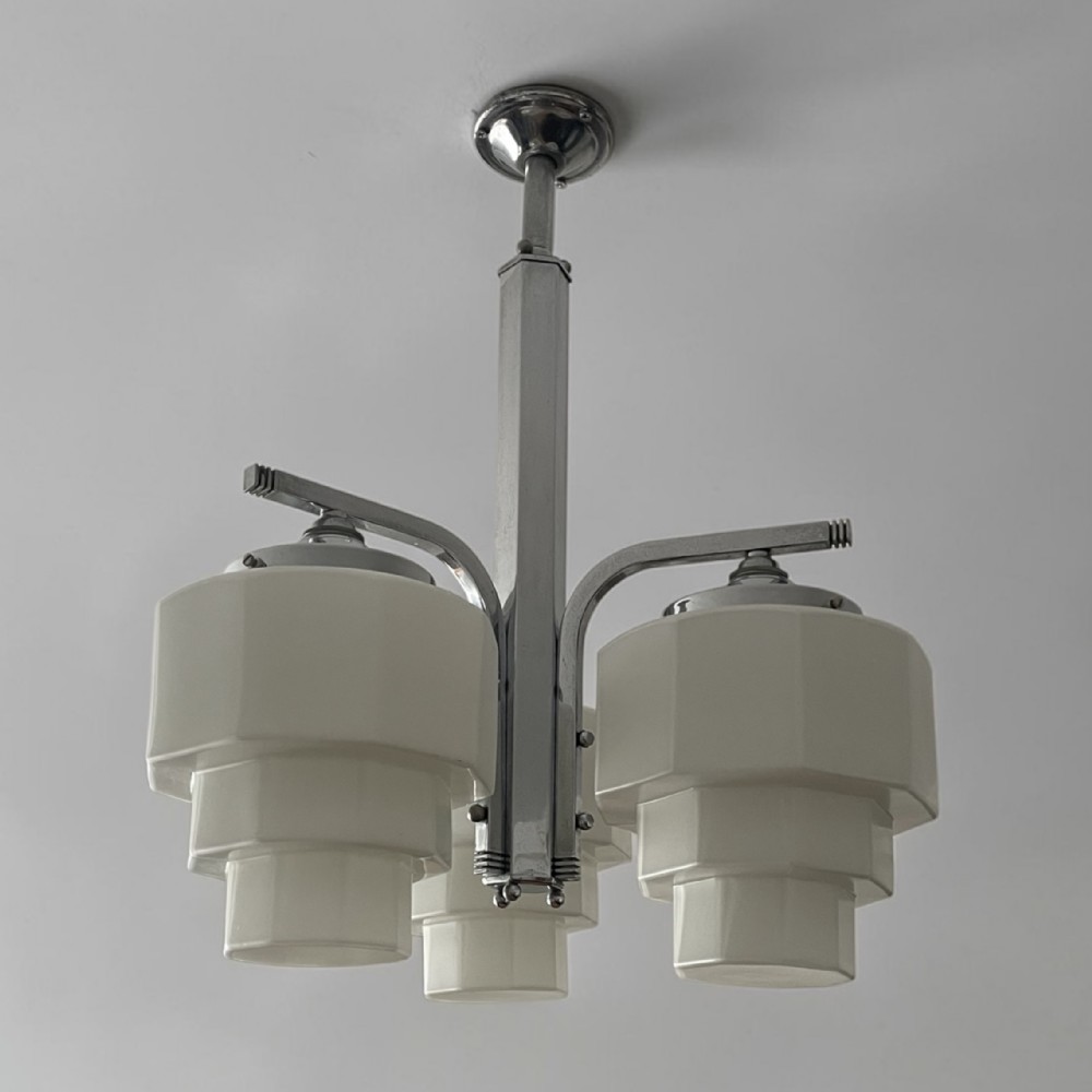 An Art Deco three-arm ceiling light fixture featuring a central chrome stem and three stepped white glass shades.