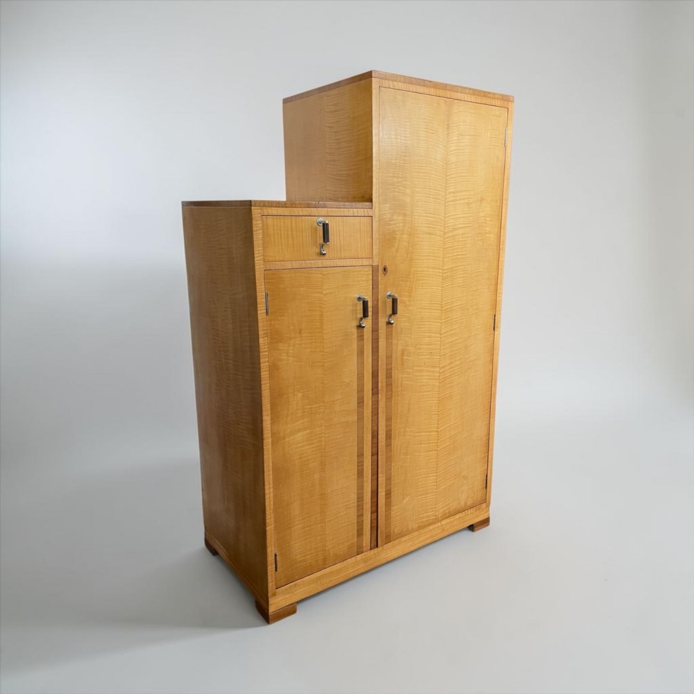 An Art Deco stepped blonde wood cabinet featuring a full-length door on the right, a smaller lower cabinet and top drawer on the left, fitted with vertical bar handles.