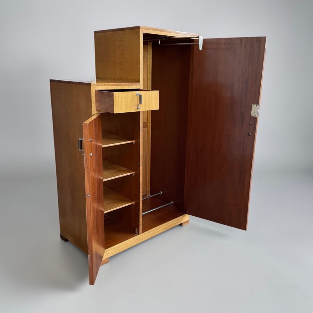 An Art Deco stepped blonde wood cabinet featuring a full-length door on the right, a smaller lower cabinet and top drawer on the left, fitted with vertical bar handles. View II