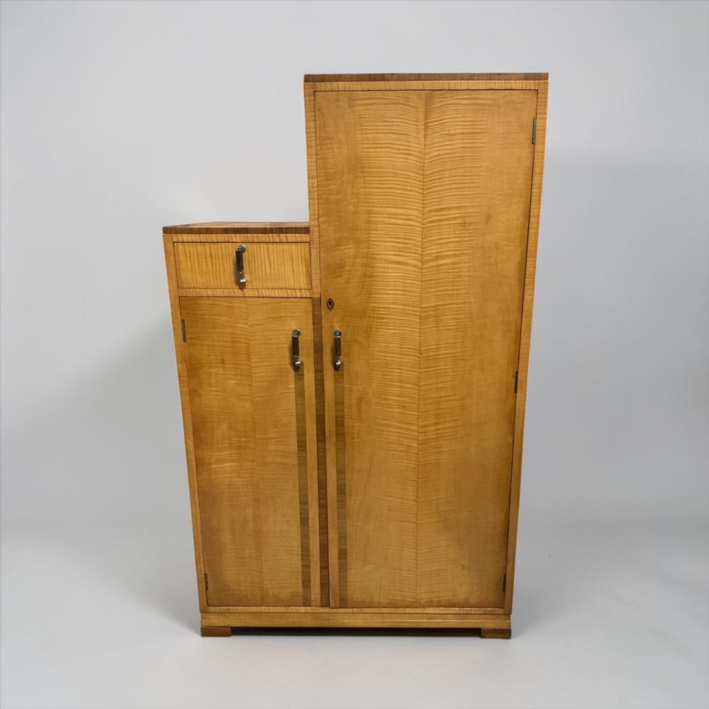 An Art Deco stepped blonde wood cabinet featuring a full-length door on the right, a smaller lower cabinet and top drawer on the left, fitted with vertical bar handles. View III