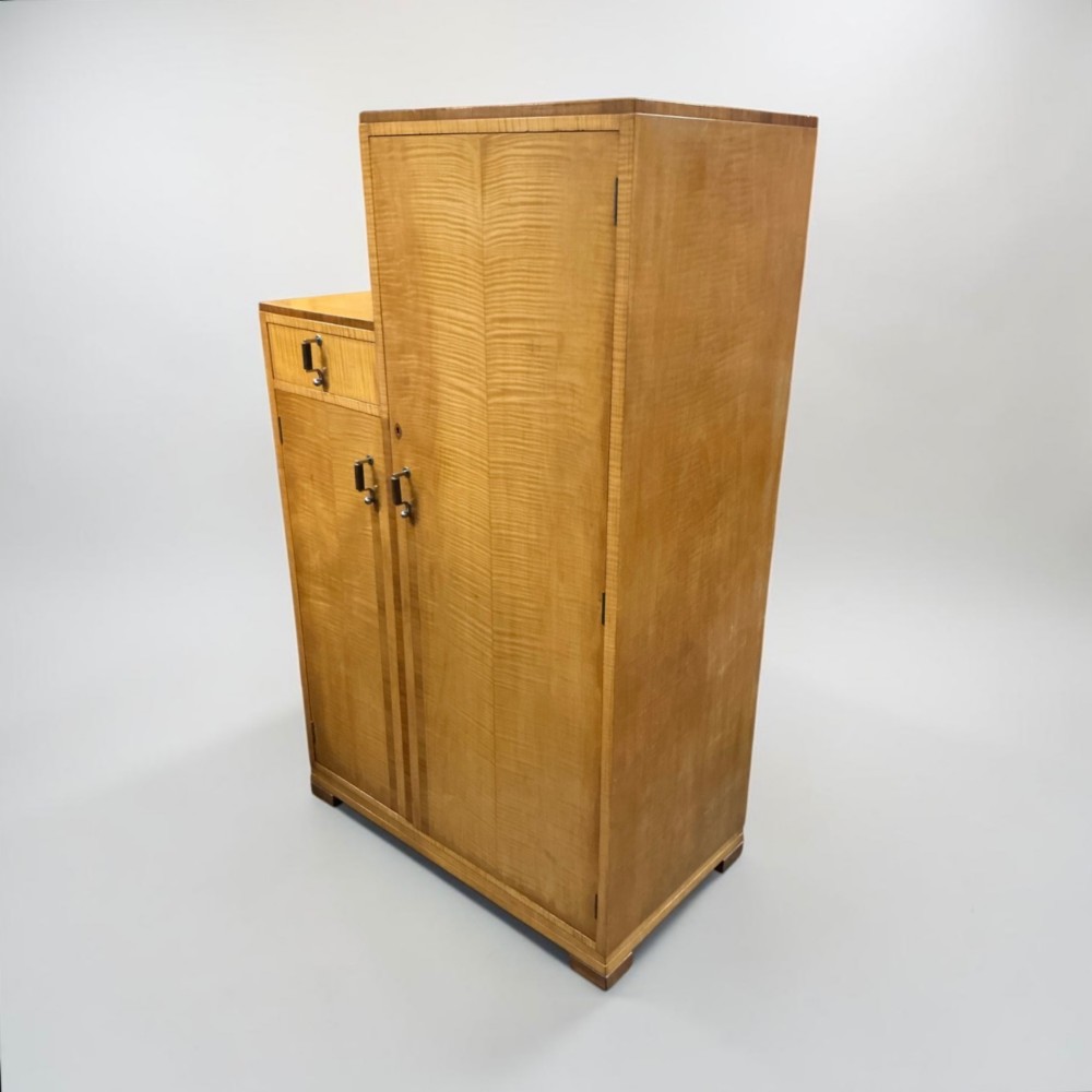 An Art Deco stepped blonde wood cabinet featuring a full-length door on the right, a smaller lower cabinet and top drawer on the left, fitted with vertical bar handles. View IV