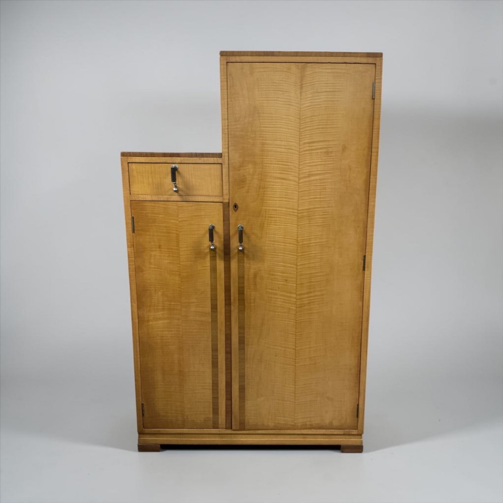 An Art Deco stepped blonde wood cabinet featuring a full-length door on the right, a smaller lower cabinet and top drawer on the left, fitted with vertical bar handles. View V