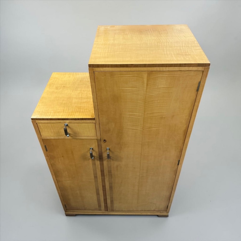 An Art Deco stepped blonde wood cabinet featuring a full-length door on the right, a smaller lower cabinet and top drawer on the left, fitted with vertical bar handles. View VI