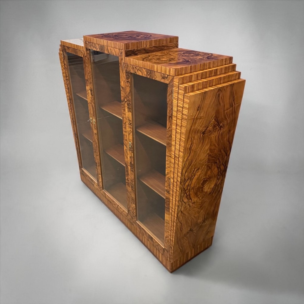 An Art Deco warm brown walnut bookcase featuring a stepped architectural top, three glass doors, and highly figured timber veneer.
