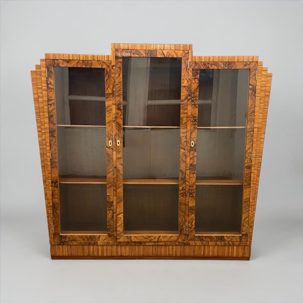 An Art Deco warm brown walnut bookcase featuring a stepped architectural top, three glass doors, and highly figured timber veneer. View III