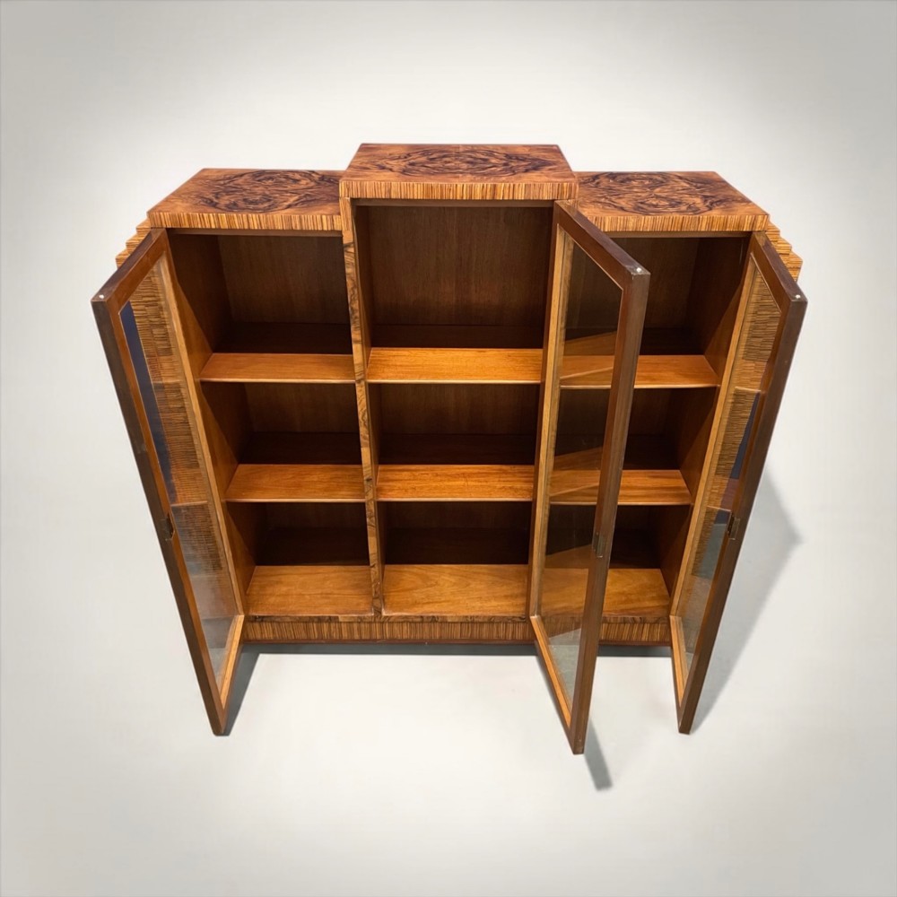 An Art Deco warm brown walnut bookcase featuring a stepped architectural top, three glass doors, and highly figured timber veneer. View V