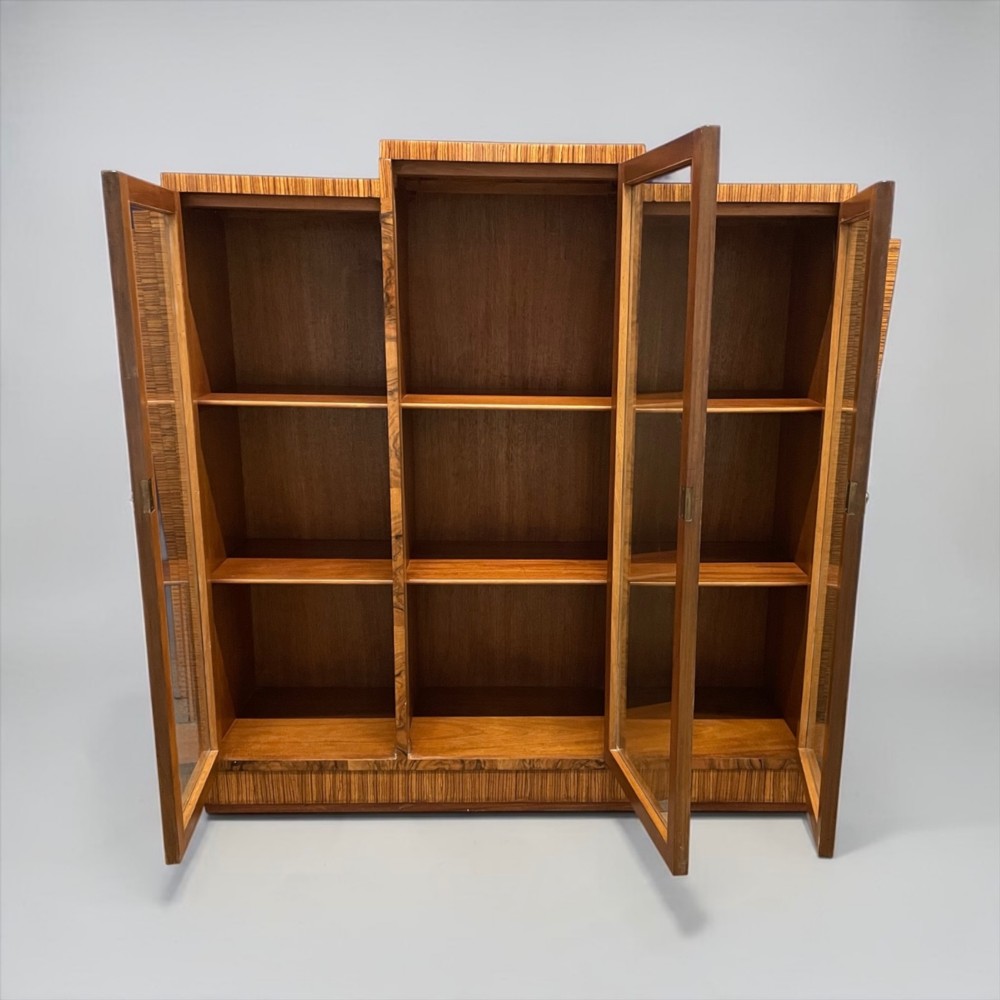 An Art Deco warm brown walnut bookcase featuring a stepped architectural top, three glass doors, and highly figured timber veneer. View VI