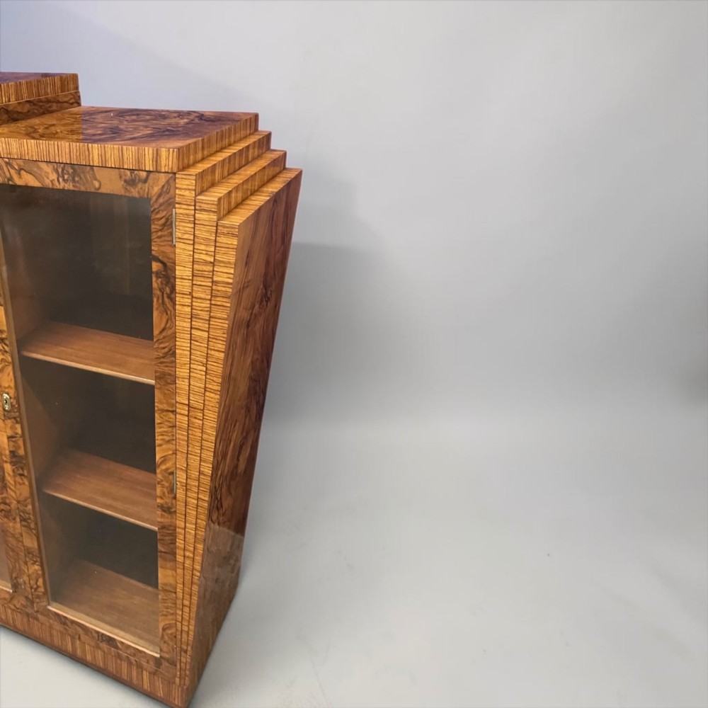 An Art Deco warm brown walnut bookcase featuring a stepped architectural top, three glass doors, and highly figured timber veneer. View VII