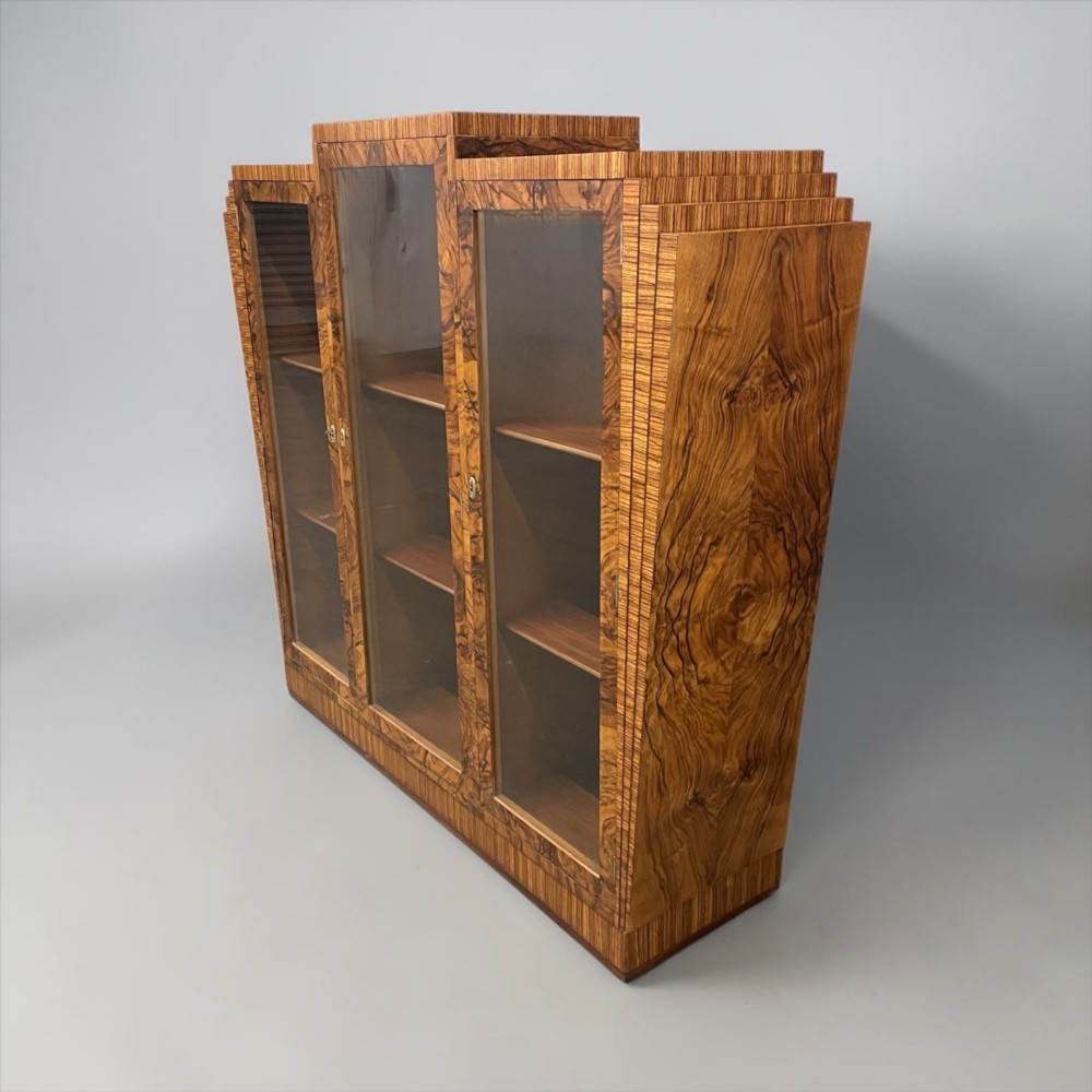 An Art Deco warm brown walnut bookcase featuring a stepped architectural top, three glass doors, and highly figured timber veneer. View X
