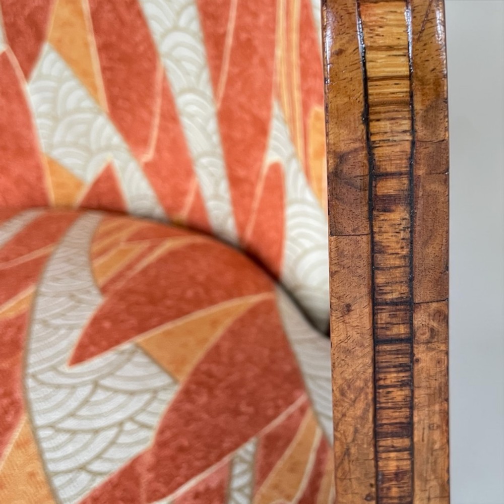 An Art Deco armchair featuring a polished walnut wooden frame, geometric orange patterned upholstery, angular armrests, and tapered legs. View X