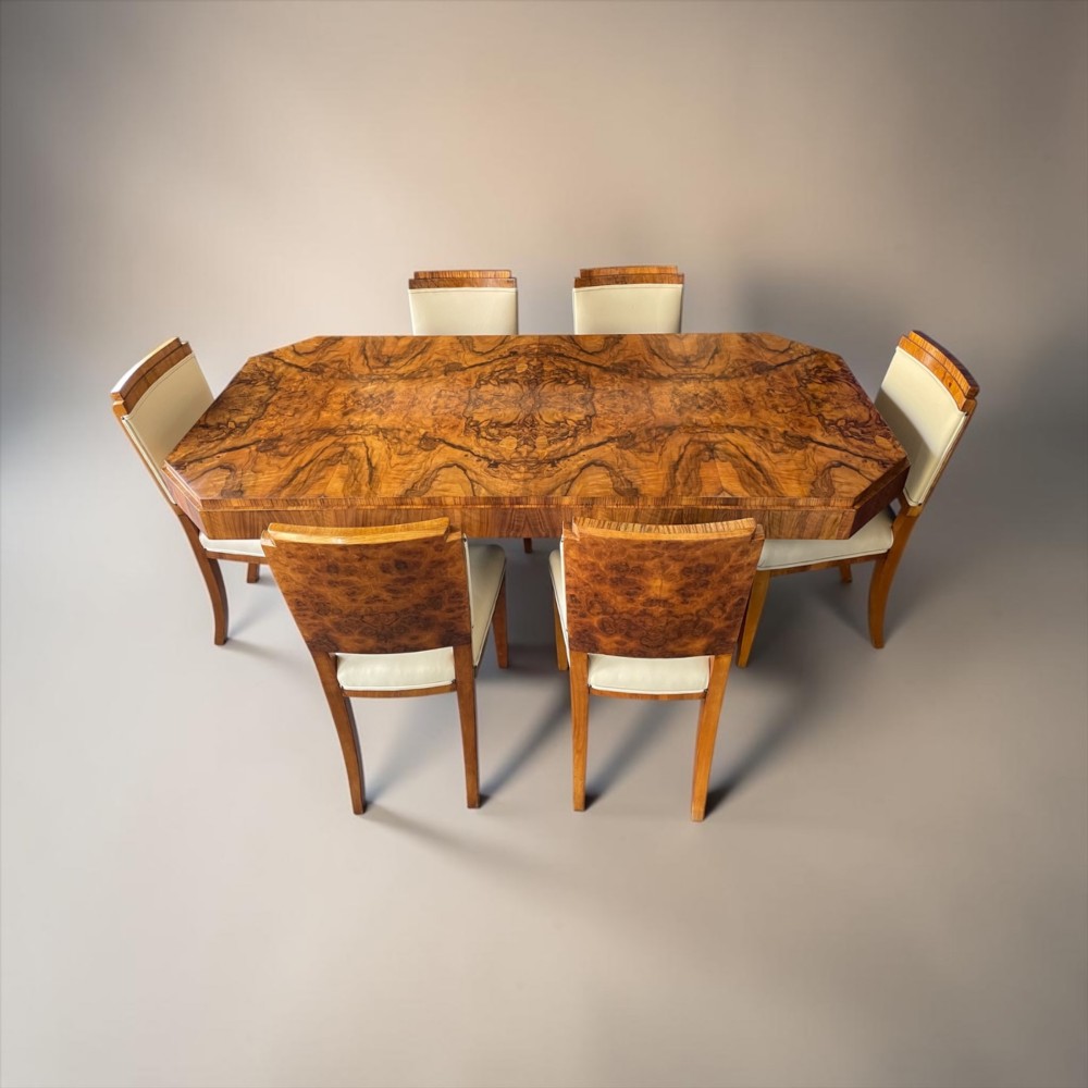 An Art Deco burr walnut dining table featuring a richly figured canted top paired with six matching chairs upholstered in cream leather.