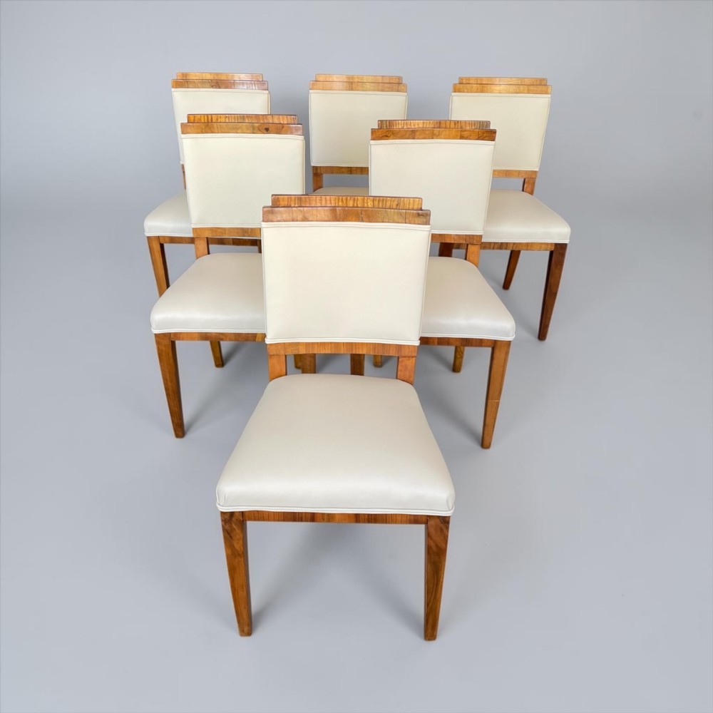 An Art Deco burr walnut dining table featuring a richly figured canted top paired with six matching chairs upholstered in cream leather. View IX