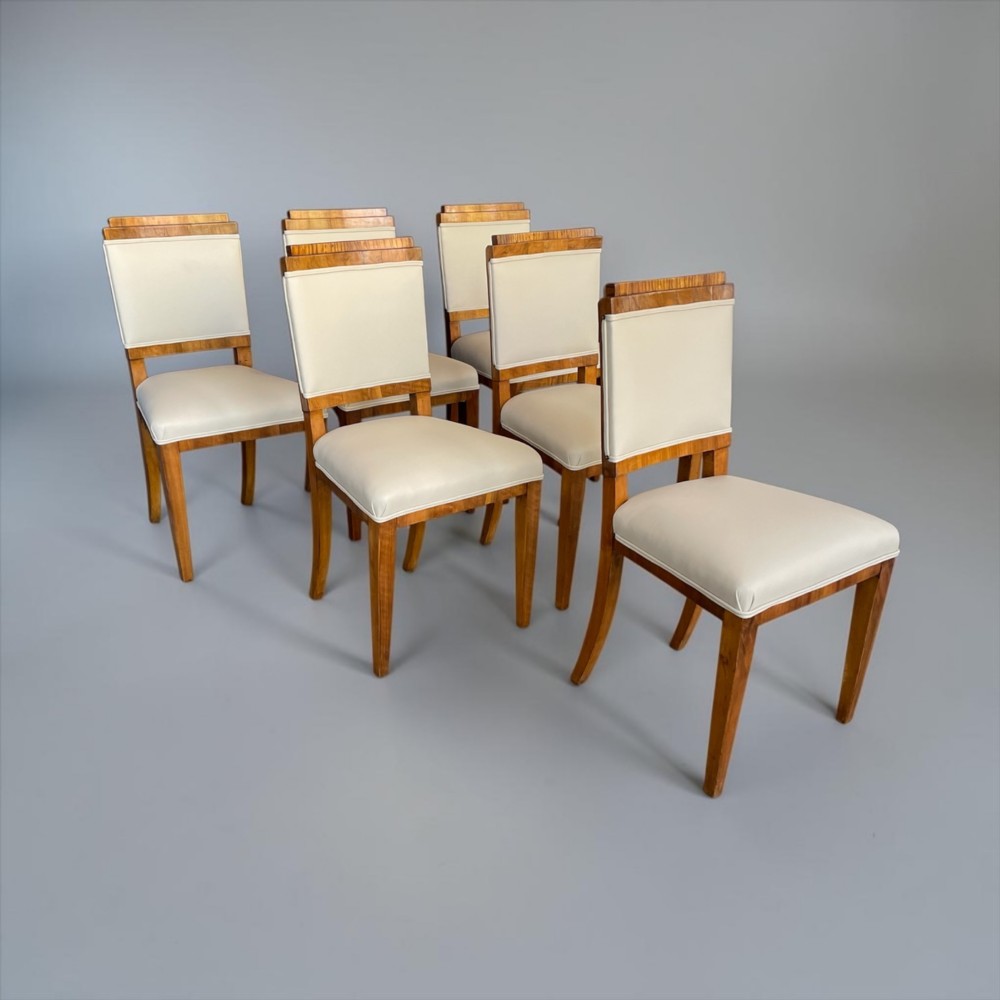 An Art Deco burr walnut dining table featuring a richly figured canted top paired with six matching chairs upholstered in cream leather. View X