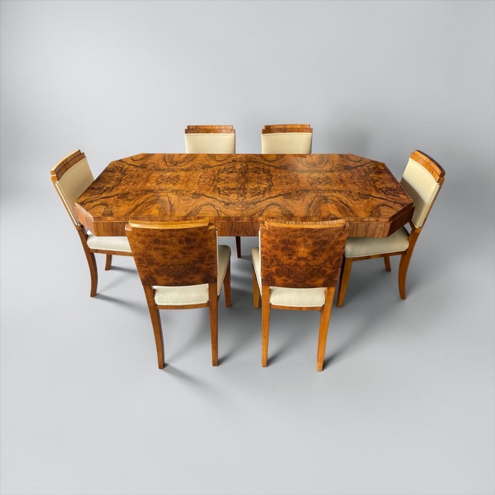 An Art Deco burr walnut dining table featuring a richly figured canted top paired with six matching chairs upholstered in cream leather. View XV