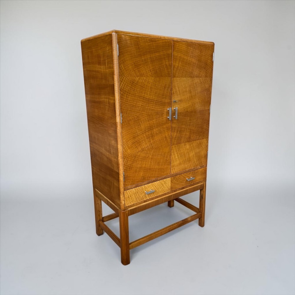 An Art Deco golden oak cabinet on stand featuring book-matched chevron wood veneers, two lower drawers, twin cupboard doors, and metal handles.