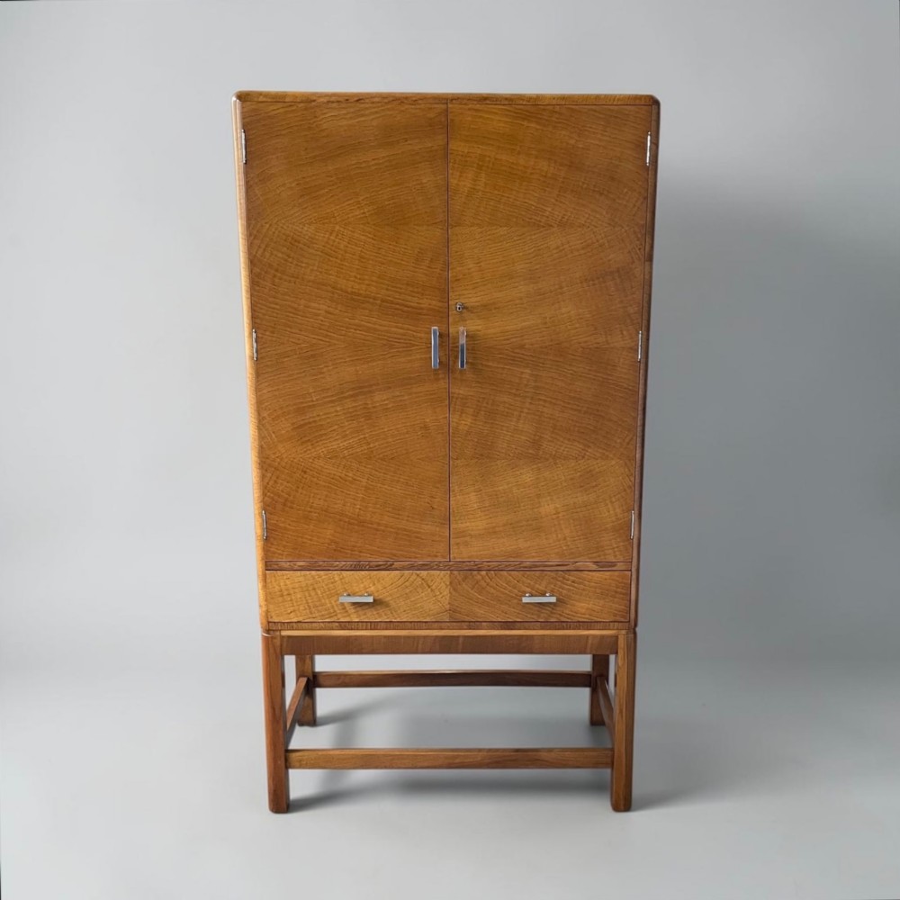 An Art Deco golden oak cabinet on stand featuring book-matched chevron wood veneers, two lower drawers, twin cupboard doors, and metal handles. View X