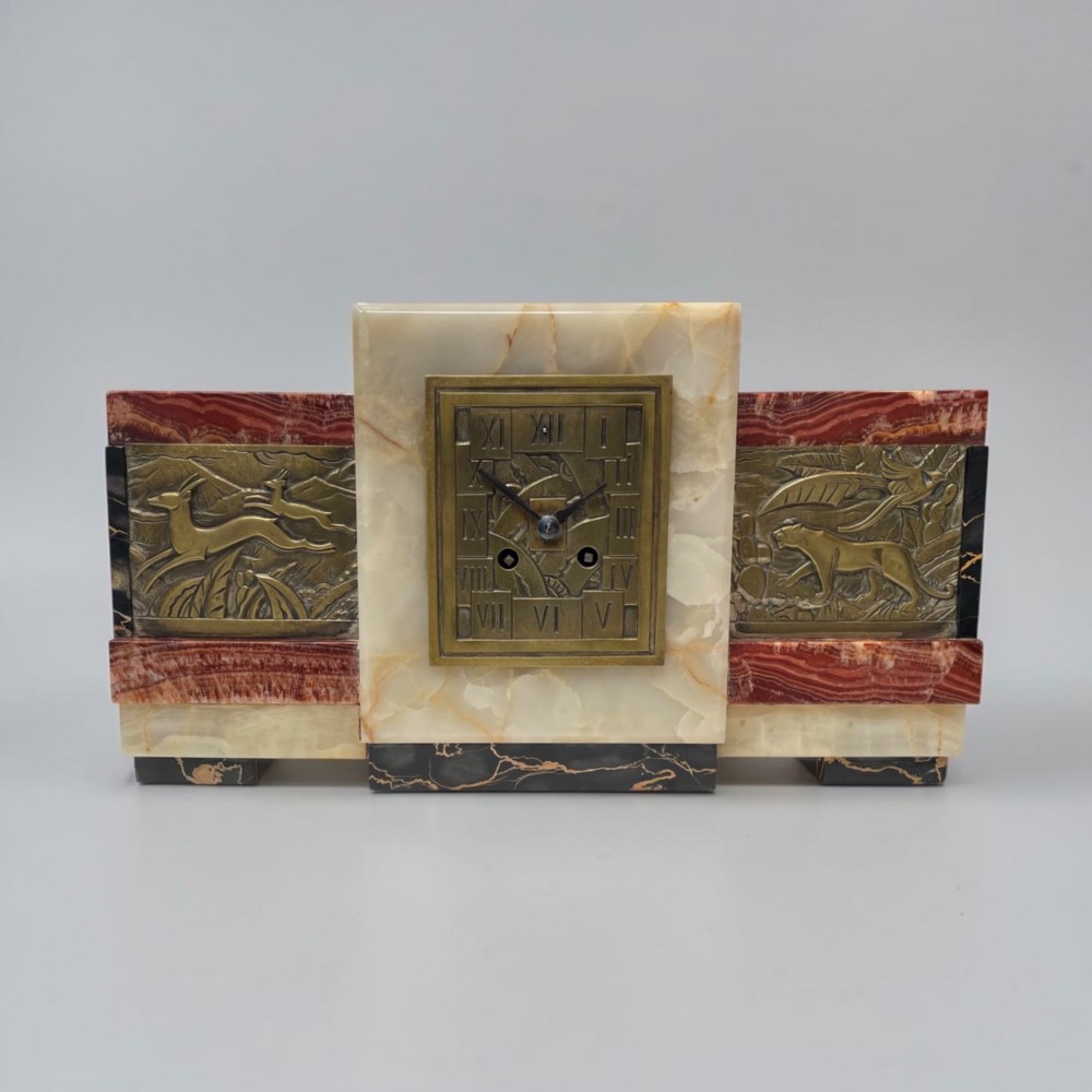 An Art Deco stepped mantel clock crafted from cream onyx and red marble with relief bronze panels depicting gazelles and a panther.