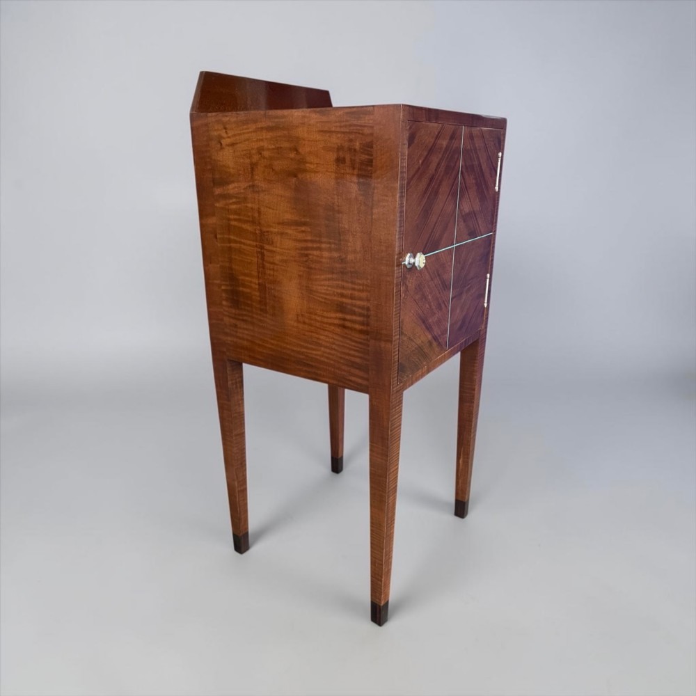 An Art Deco figured walnut bedside table featuring a raised gallery back, geometric parquetry double doors, and slender tapered legs.