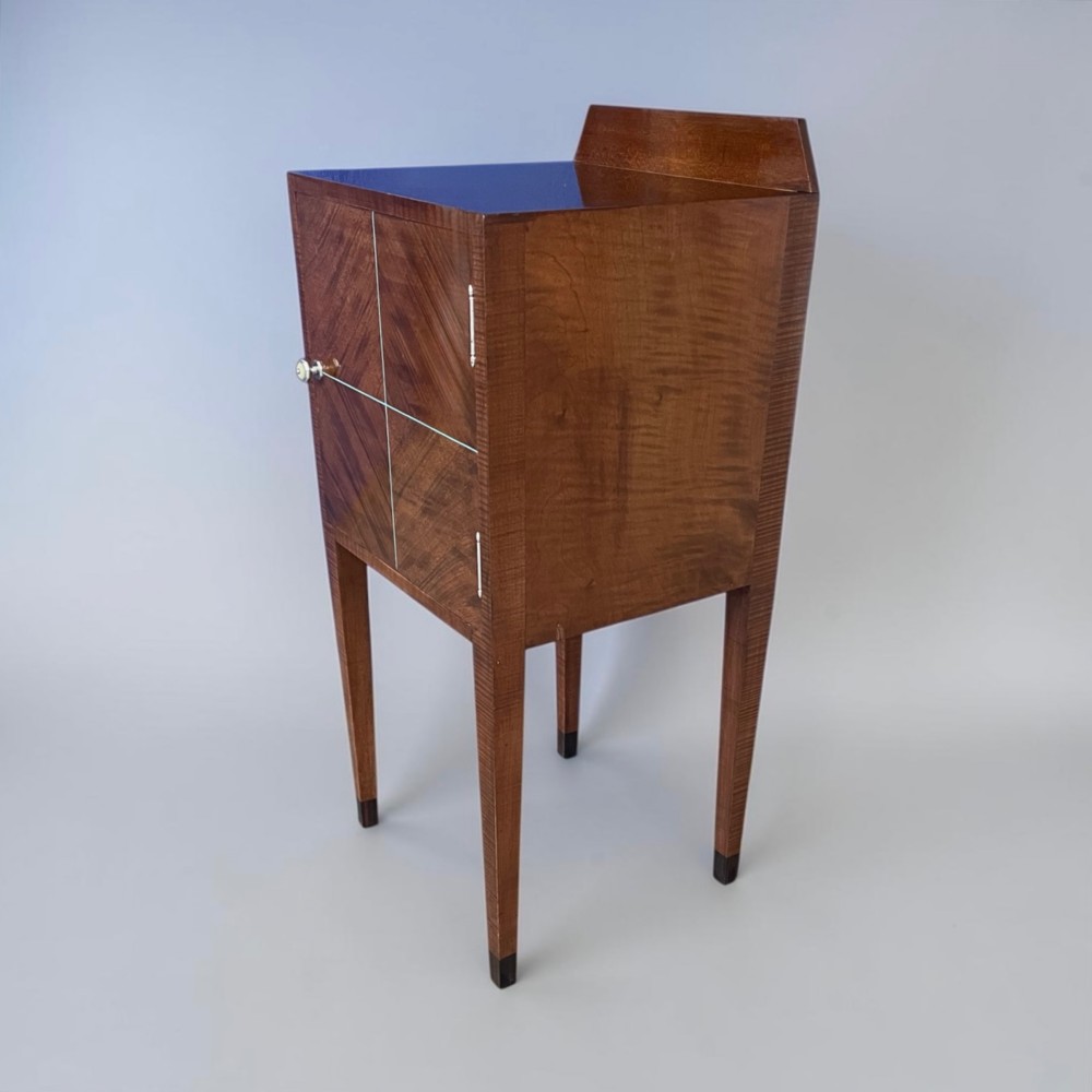 An Art Deco figured walnut bedside table featuring a raised gallery back, geometric parquetry double doors, and slender tapered legs. View X