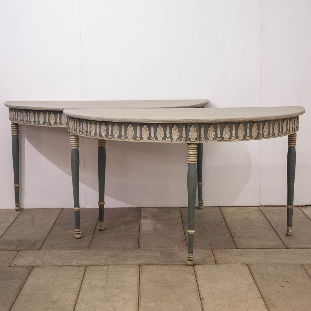 A pair of semi-circular Swedish Gustavian style console tables featuring grey and off-white painted finishes with carved leaf patterns around the apron and tapered legs.