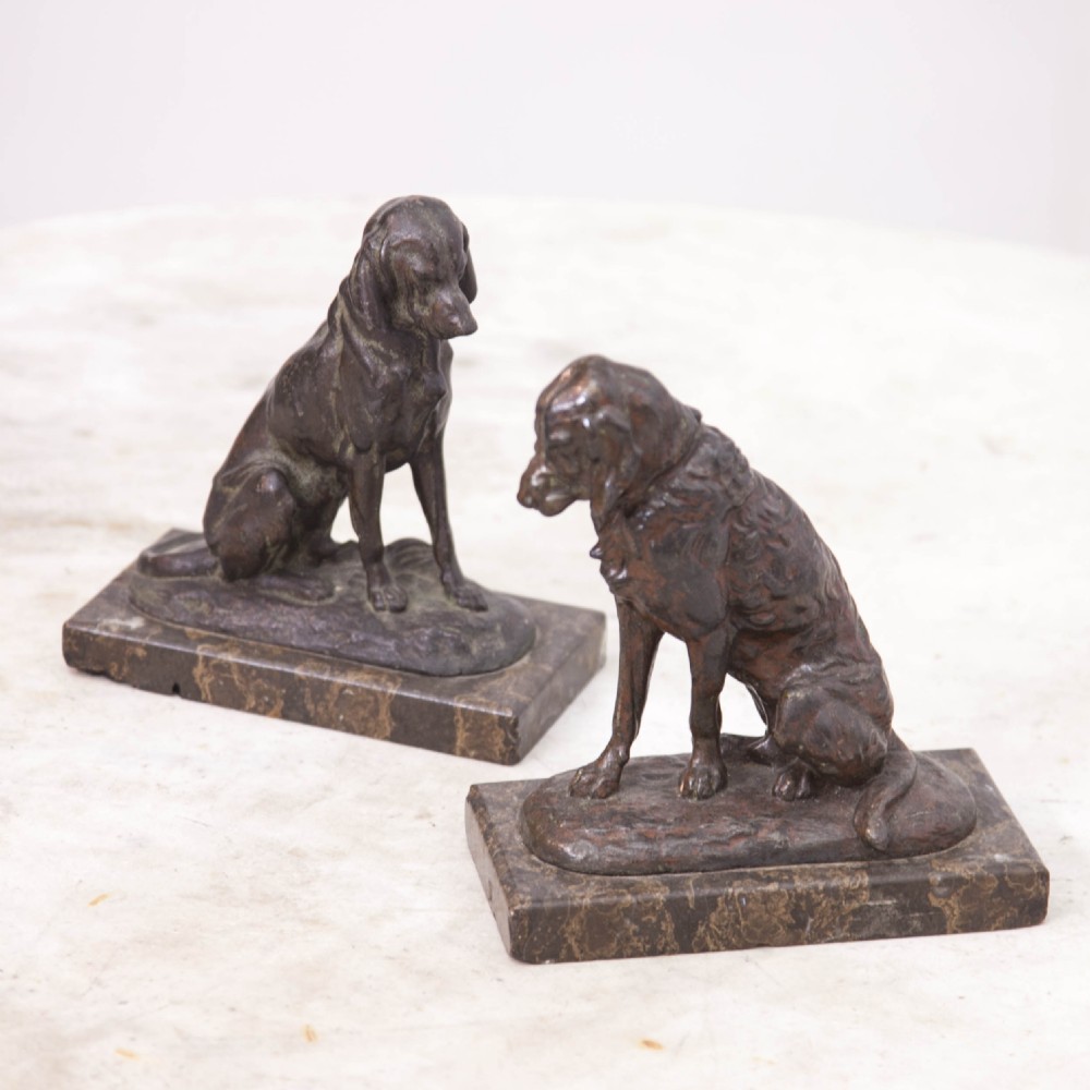 A pair of patinated cast metal figures depicting seated dogs mounted on rectangular dark marble plinths.