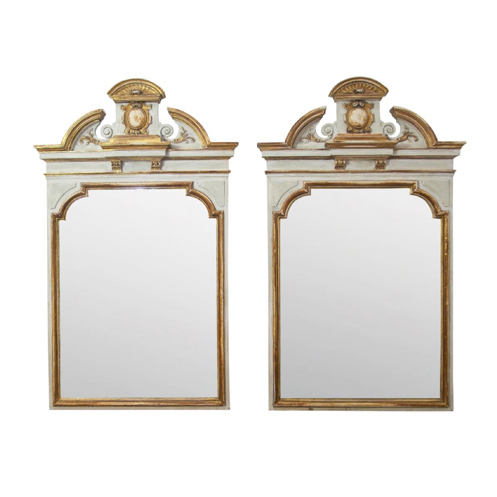 A matching pair of Neoclassical wall mirrors featuring cream painted frames with gilded pediments, scroll cresting, and rectangular mirror glass.