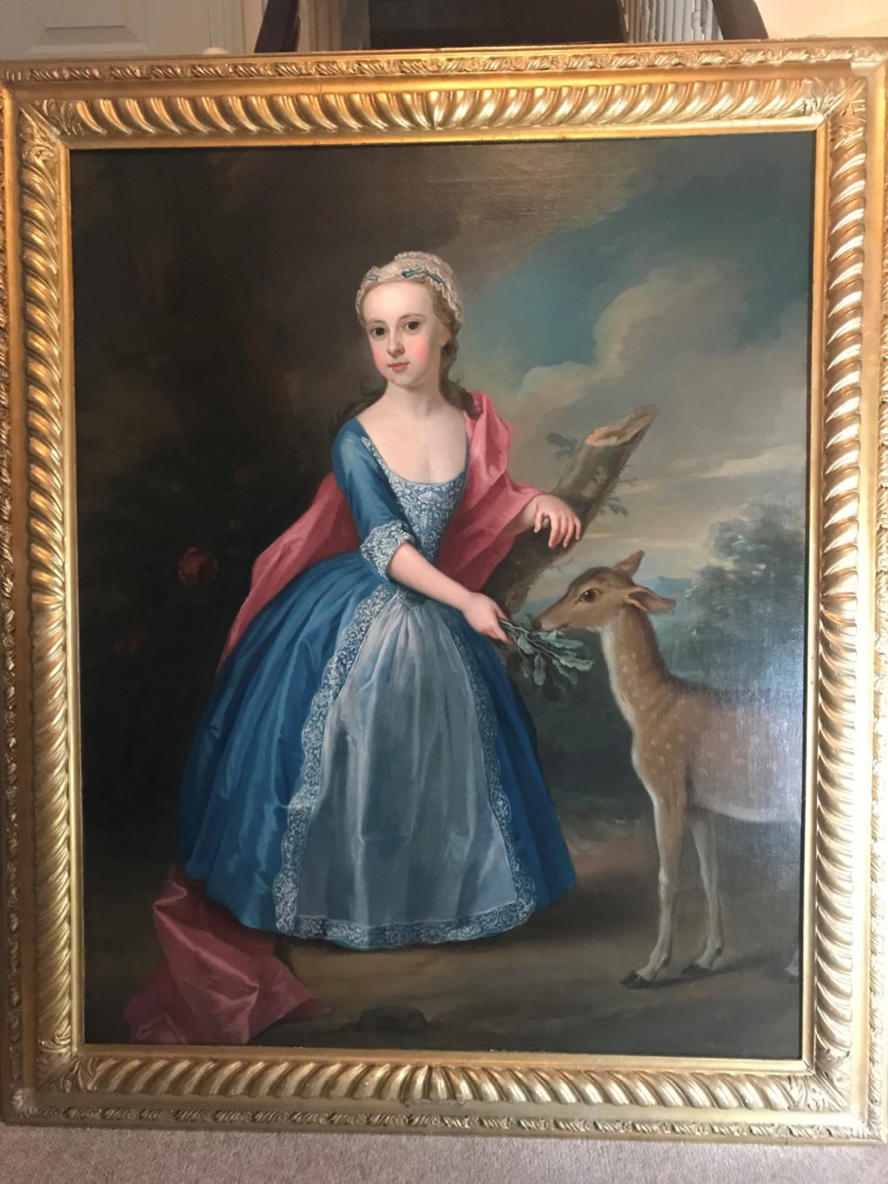 An 18th-century oil painting on canvas depicting a young girl in a blue dress and pink mantle feeding leaves to a deer, set within a carved giltwood frame.