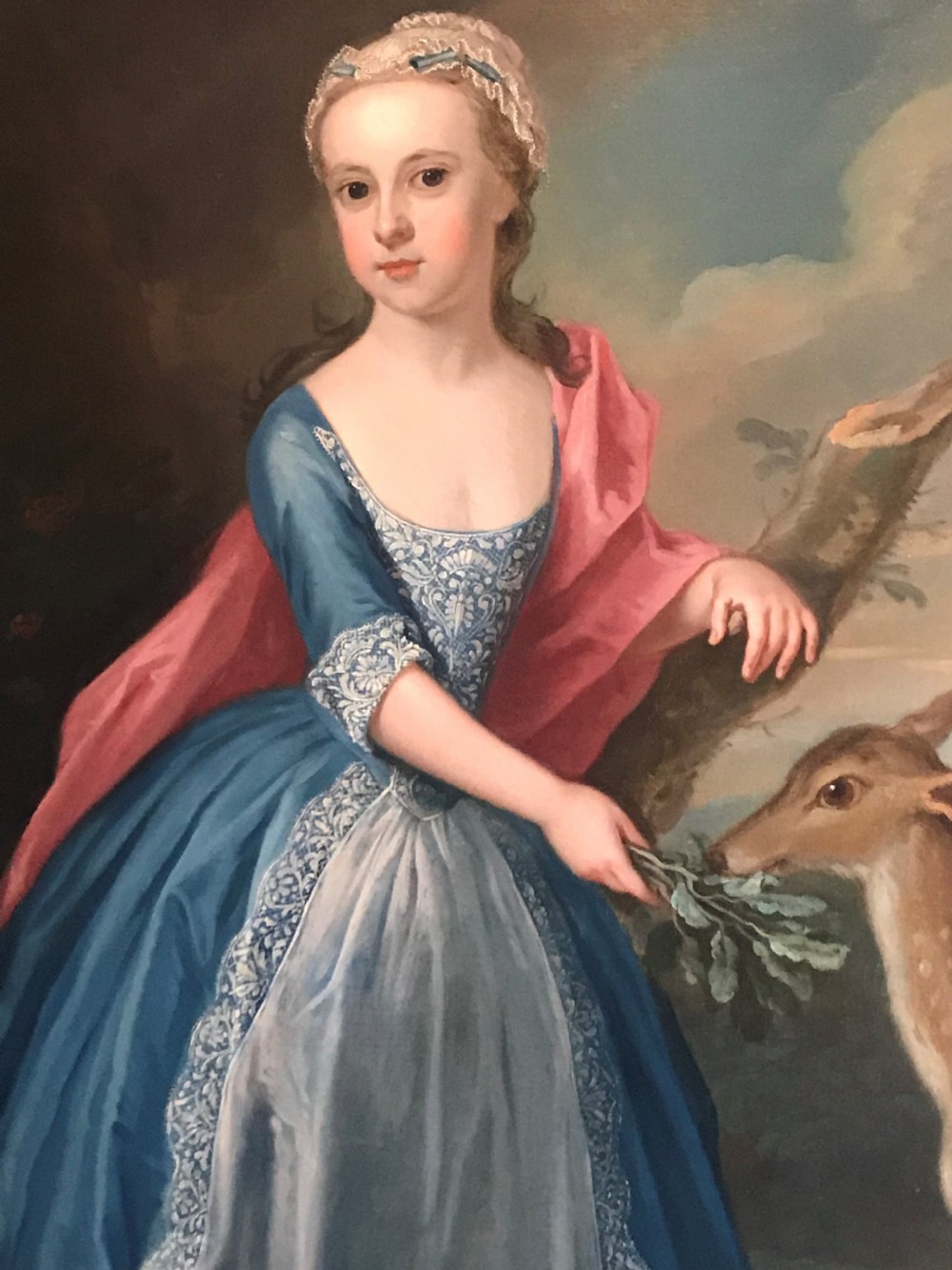 An 18th-century oil painting on canvas depicting a young girl in a blue dress and pink mantle feeding leaves to a deer, set within a carved giltwood frame. View II