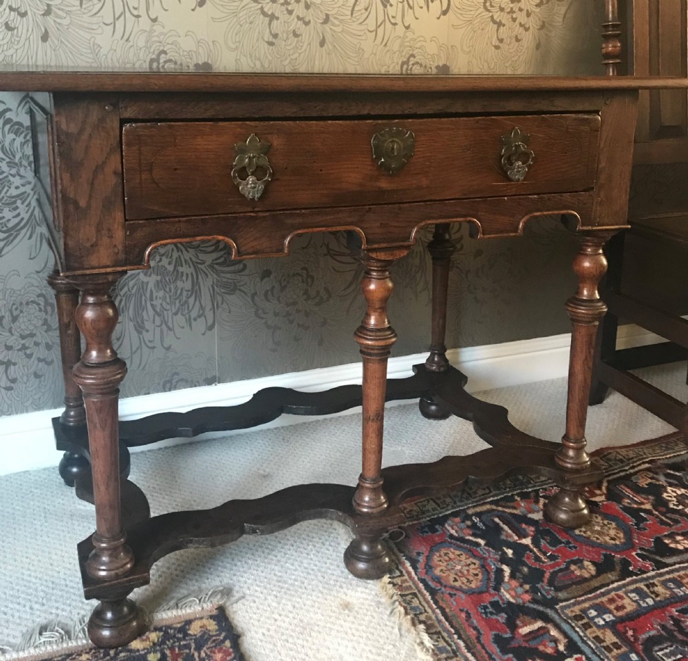 A dark oak side table with a single drawer featuring brass drop handles, a shaped apron, turned baluster legs, and a wavy stretcher base.