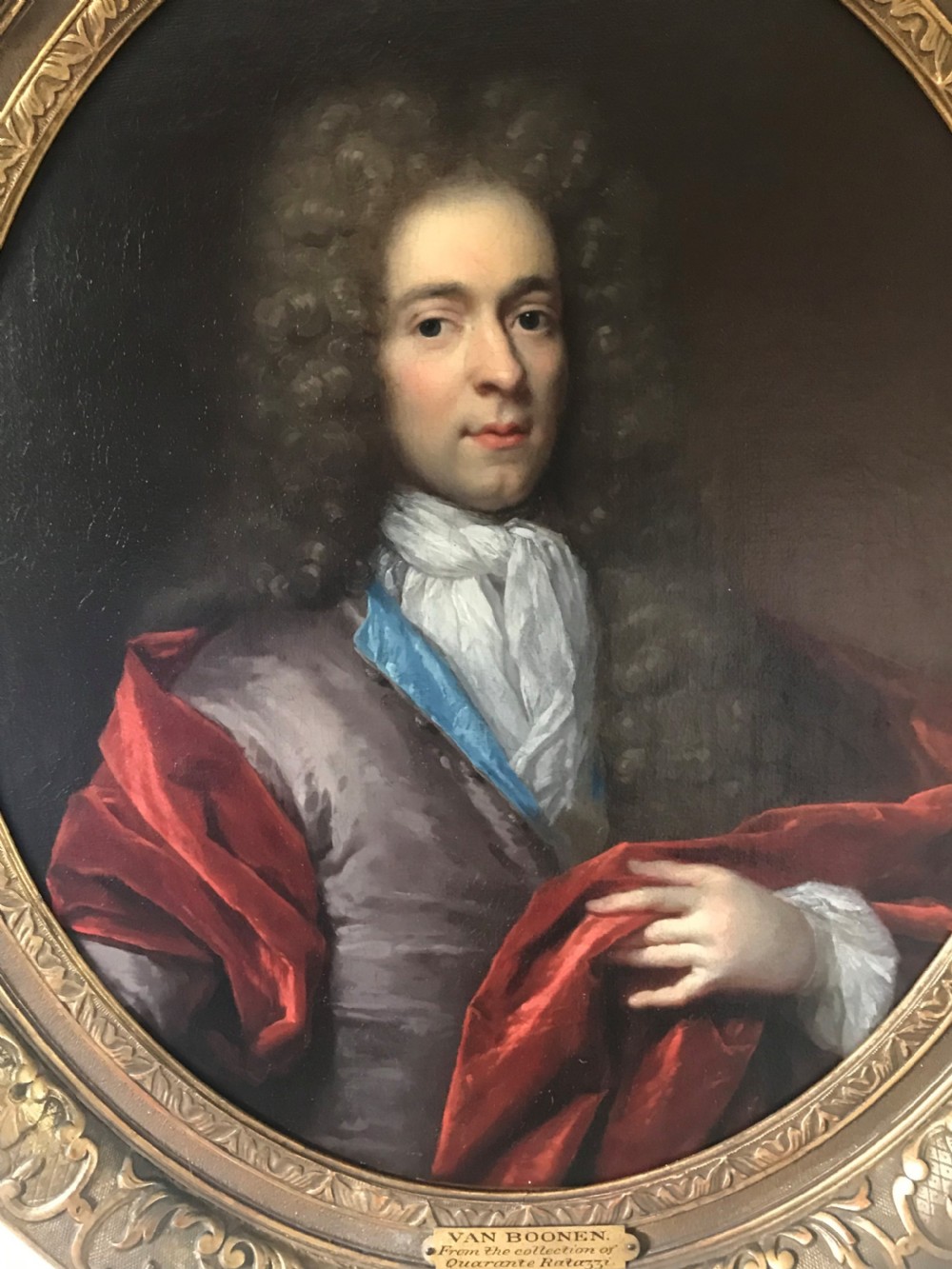 An oval seventeenth-century oil painting depicting a gentleman wearing a periwig and crimson cloak, set inside a carved giltwood frame. View II