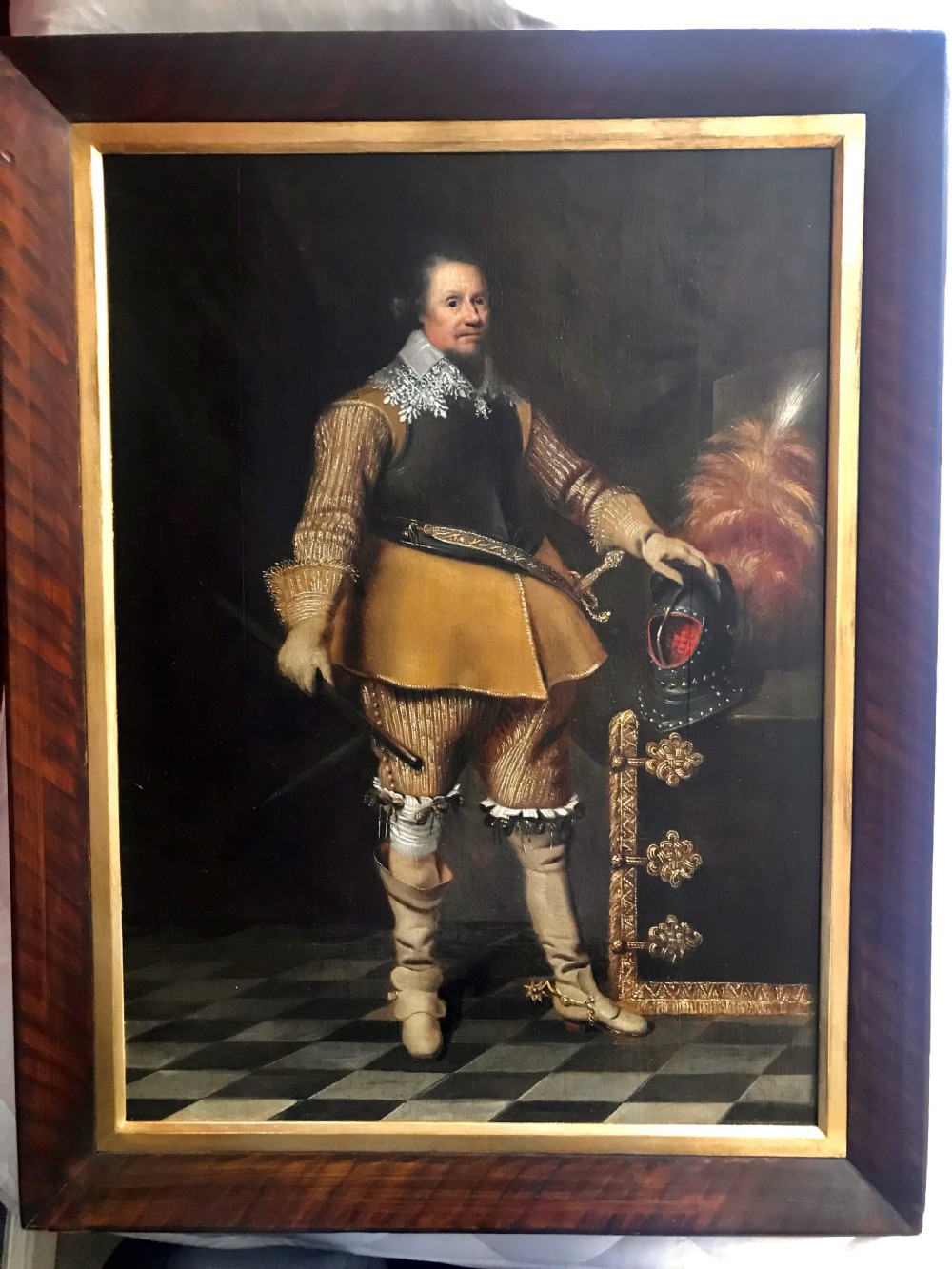 A full-length oil portrait depicting a 17th-century military commander standing beside a plumed helmet, set within a dark timber frame with a gold slip.