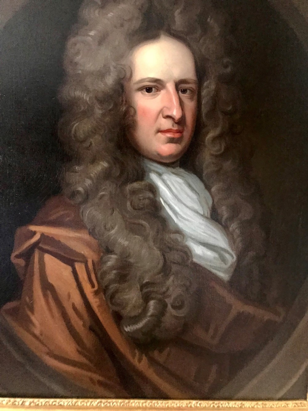 A seventeenth-century oil portrait depicting a gentleman with a large curling periwig and white cravat set inside a feigned oval within a gilded frame. View II