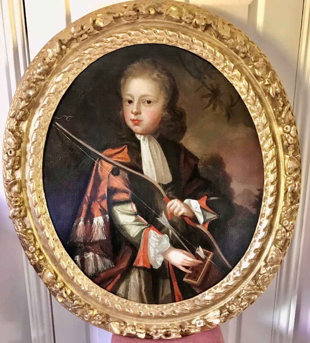 An oval Baroque oil painting depicting a young boy holding a bow, set within an ornate carved giltwood frame.