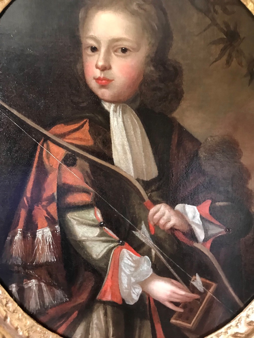 An oval Baroque oil painting depicting a young boy holding a bow, set within an ornate carved giltwood frame. View II