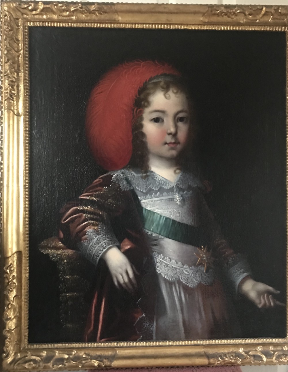 A seventeenth-century Baroque oil painting on canvas depicting a young child wearing a crimson feathered hat and ornate lace attire within a gilded wood frame.