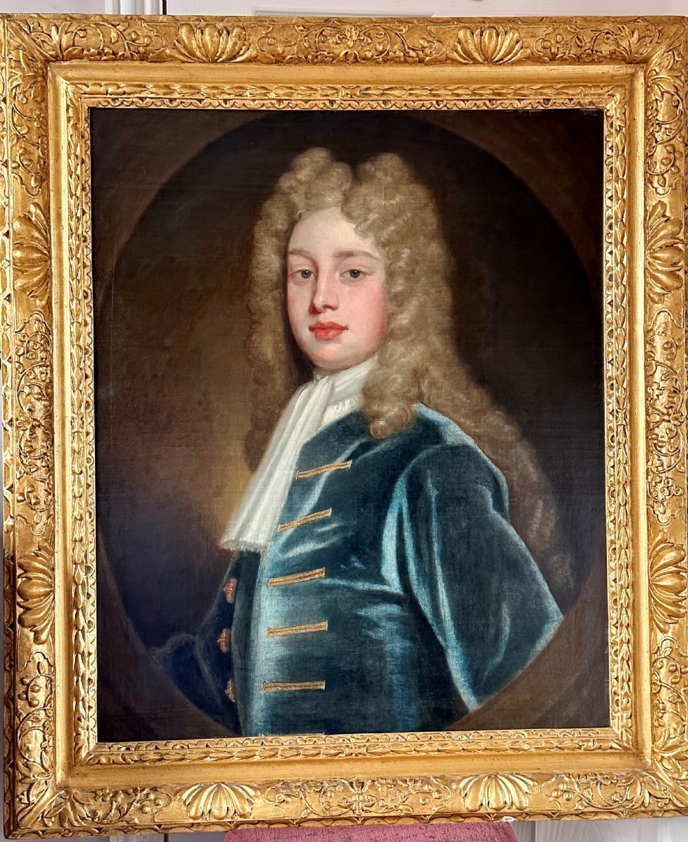 An 18th-century oil portrait painting depicting a gentleman with a long wig, white cravat, and teal blue coat, set within a carved gold frame.