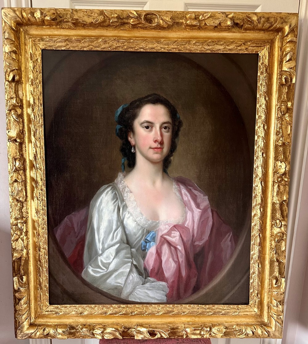 An 18th-century oil portrait of a woman in a pink and white dress set within a feigned oval and enclosed by a gold carved giltwood frame.