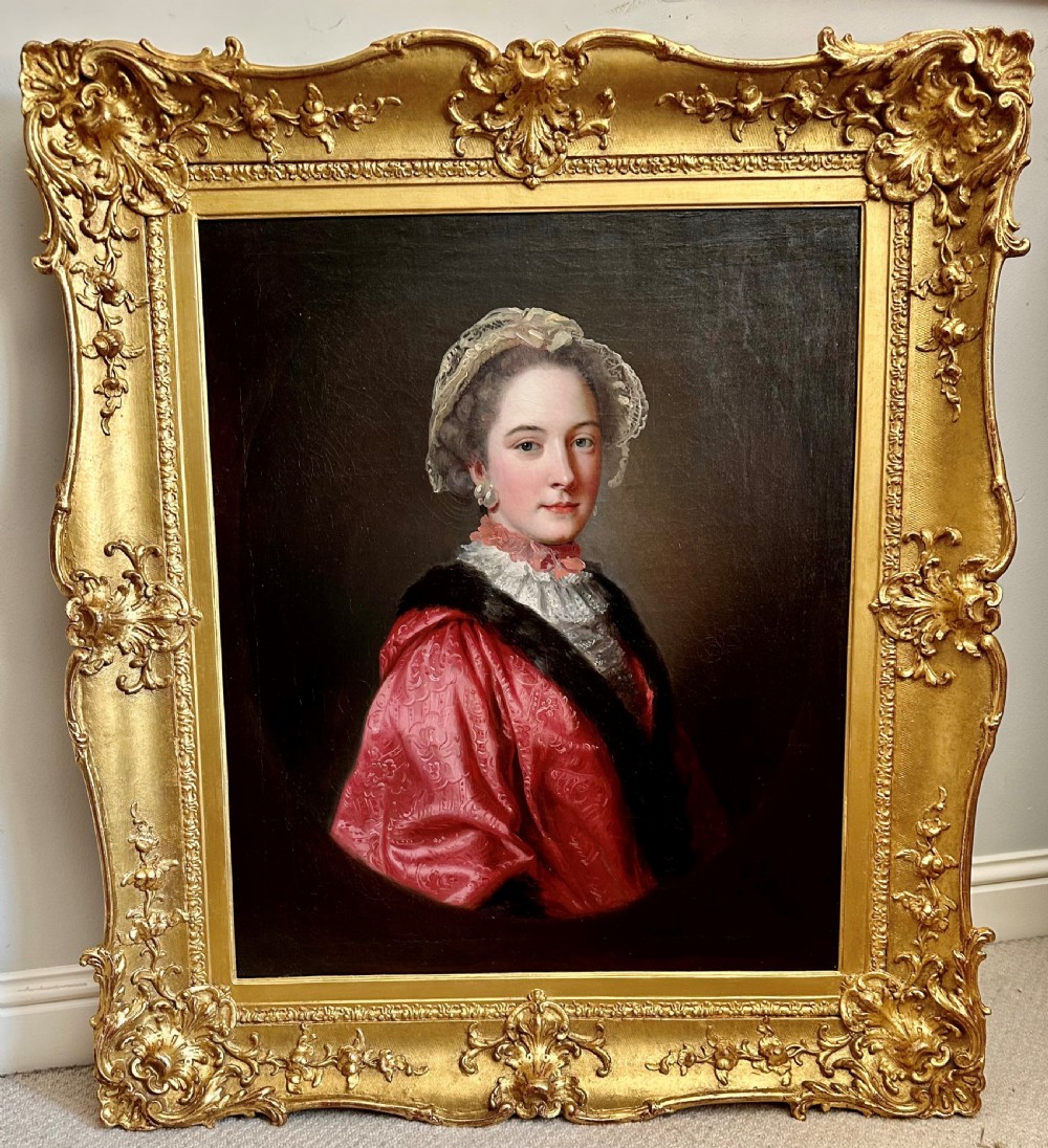 An 18th-century oil painting of a lady wearing a pink embroidered dress and lace bonnet, set within a carved gold swept frame.