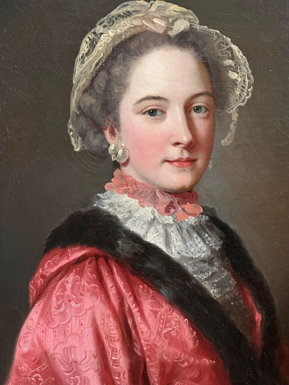 An 18th-century oil painting of a lady wearing a pink embroidered dress and lace bonnet, set within a carved gold swept frame. View II
