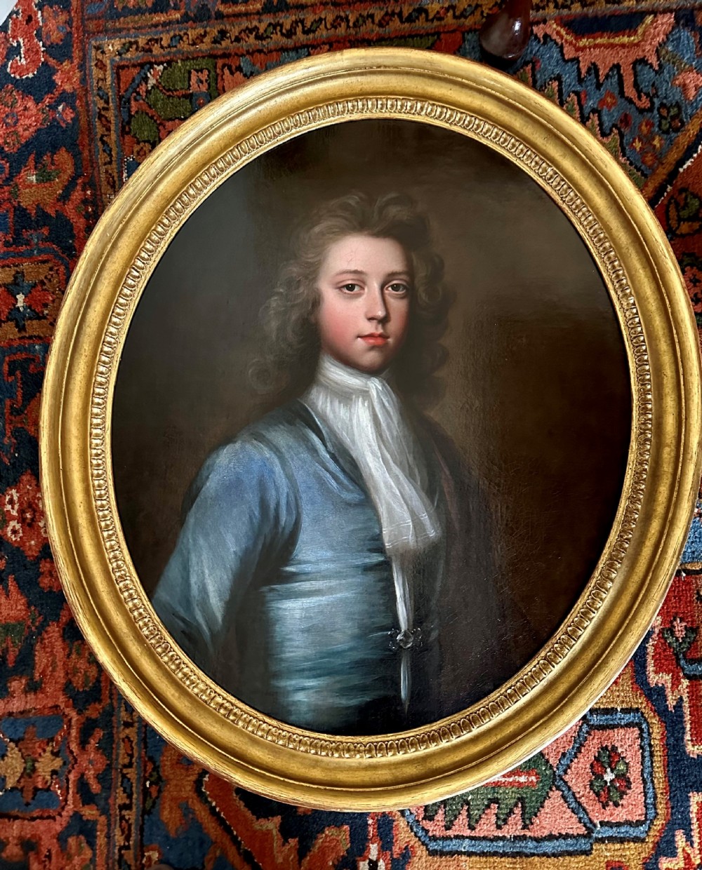 An oval 18th-century oil painting depicting a young gentleman wearing a blue coat and white cravat, set inside a carved giltwood frame.