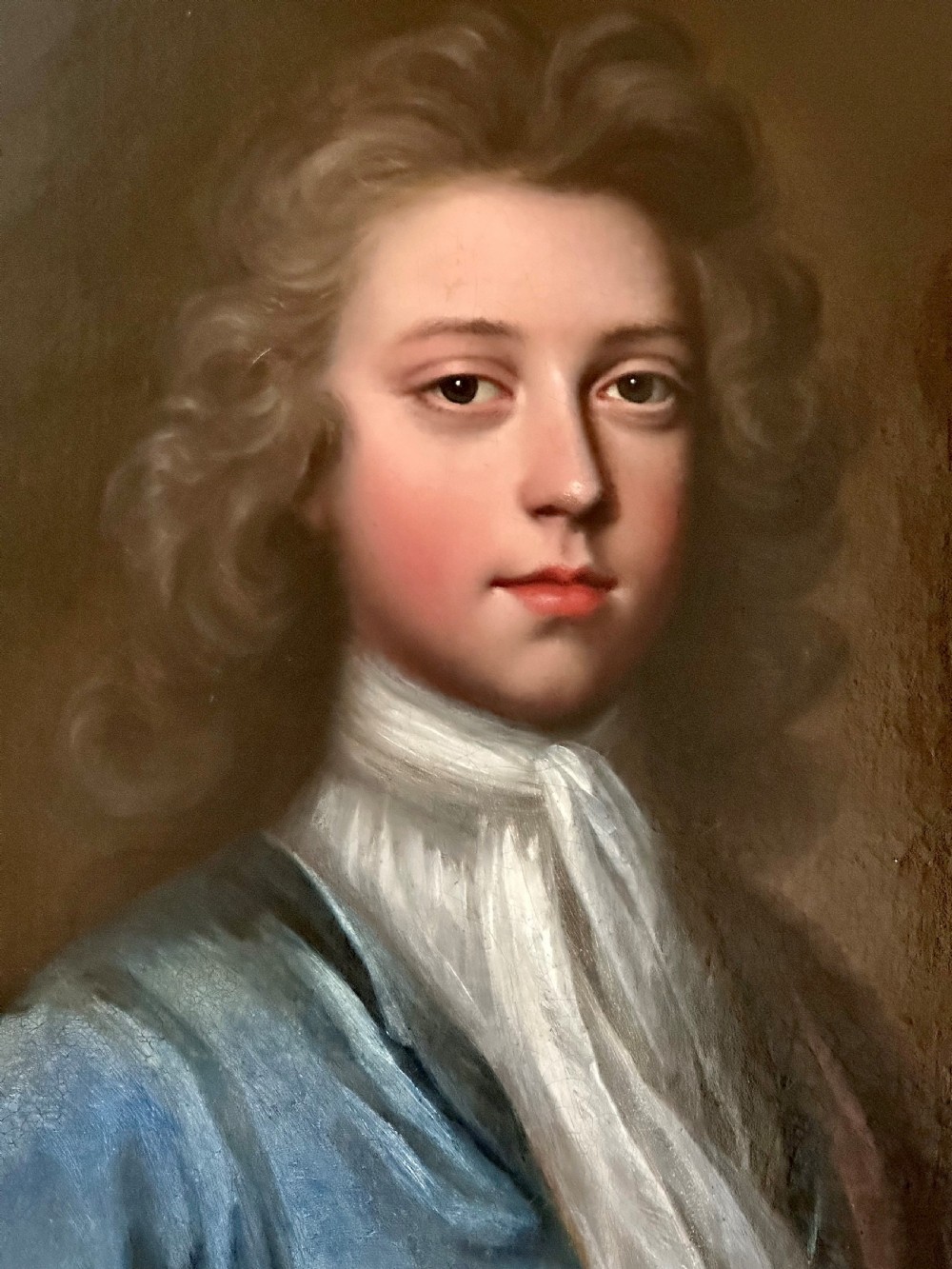 An oval 18th-century oil painting depicting a young gentleman wearing a blue coat and white cravat, set inside a carved giltwood frame. View II