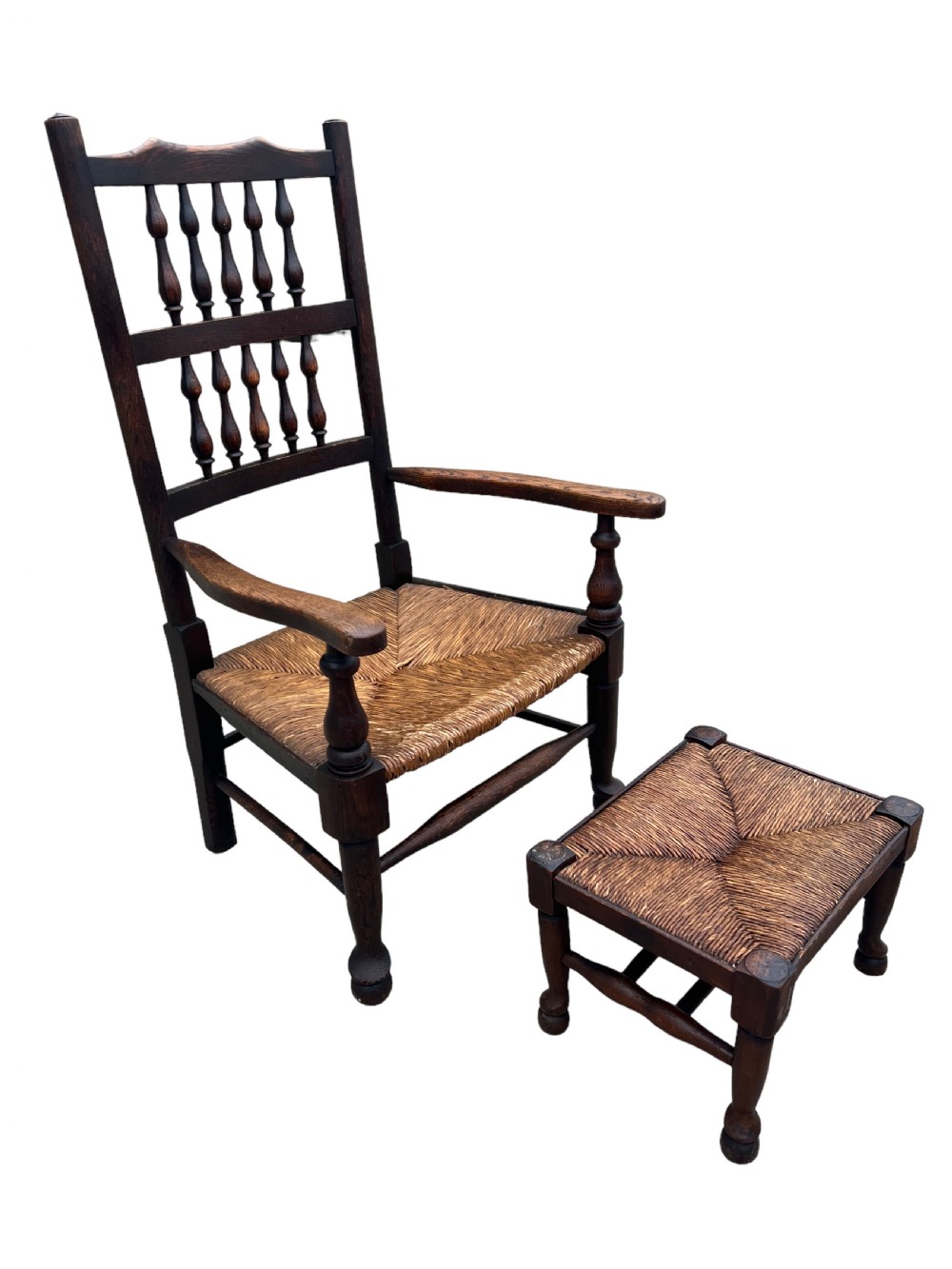 An antique dark oak armchair featuring a double-tiered turned spindle back, curved armrests, and a woven rush seat, accompanied by a matching square rush-seated footstool. View II