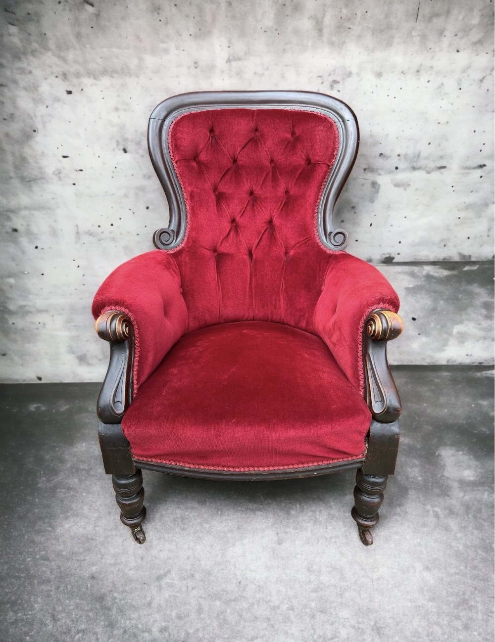 A Victorian spoon-back armchair upholstered in crimson red button-tufted velvet with a dark carved timber frame and turned front legs on castors.